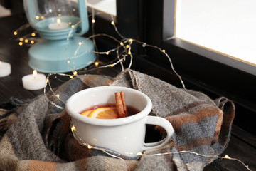 Composition with cup of hot winter drink, scarf and Christmas lights near window. Cozy season