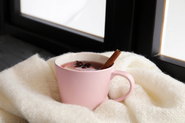 Cup of hot winter drink with knitted scarf near window. Cozy season