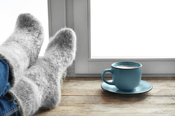 Woman in warm socks and cup of hot winter drink on windowsill. Cozy season