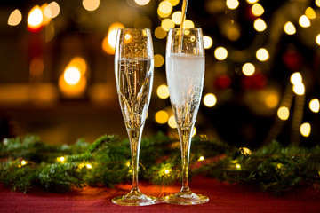 Background of two glasses of champagne, spruce branches on red table cloth. Living room decorated with lights and candles and Christmas tree