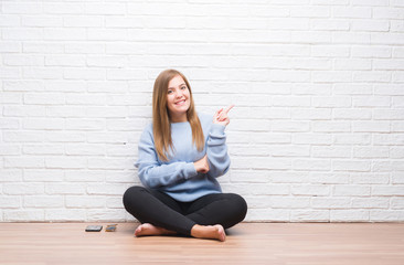 Young adult woman sitting on the floor in autumn over white brick wall with a big smile on face, pointing with hand and finger to the side looking at the camera.