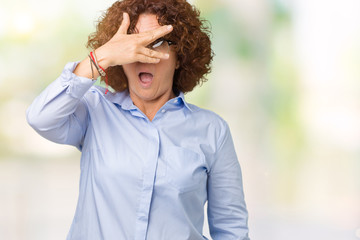 Beautiful middle ager senior businees woman wearing glasses over isolated background peeking in shock covering face and eyes with hand, looking through fingers with embarrassed expression.