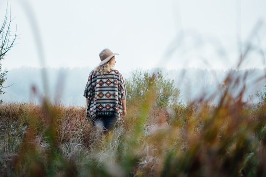 Woman in wide-brimmed felt hat and authentic poncho standing in high grass at foggy morning