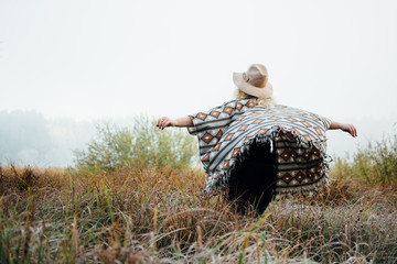 Woman in wide-brimmed felt hat and authentic poncho standing in high grass at foggy morning