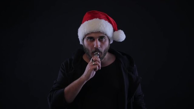 Portrait of a man with beard in Santa red hat smoking electronic cigarette. Bad addiction concept. Vape pen. Vape smoke. Christmas. Black background
