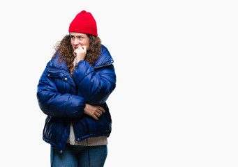 Beautiful young brunette curly hair girl wearing winter coat, wool cap and sweater over isolated background looking stressed and nervous with hands on mouth biting nails. Anxiety problem.