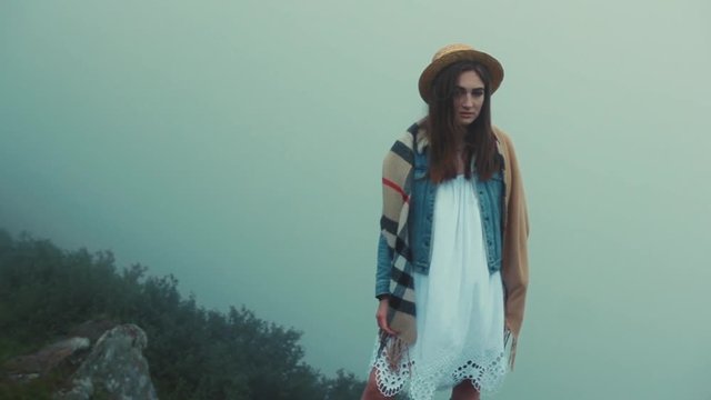 Serious young woman with hat in a white dress stand in mountains looking at camera fog beautiful nature stylish travel satisfied tourist traveler landscape sky tourism portrait close up slow motion