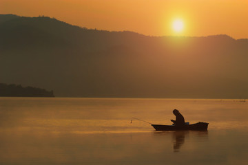 Silhouette lonely fishing man sitting in his boat and look smart phone with sunset sky and selective focus, Background for weekend travel or recreation or relax activity after working hard, Thailand.