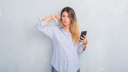 Young adult woman over grey grunge wall looking at smartphone texting a message with angry face,...
