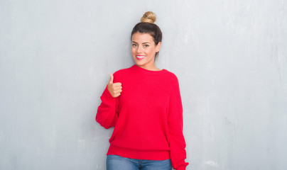 Obraz na płótnie Canvas Young adult woman over grey grunge wall wearing winter outfit doing happy thumbs up gesture with hand. Approving expression looking at the camera with showing success.