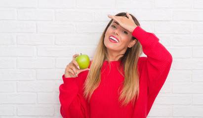 Young adult woman over white brick wall eating fresh green apple stressed with hand on head, shocked with shame and surprise face, angry and frustrated. Fear and upset for mistake.