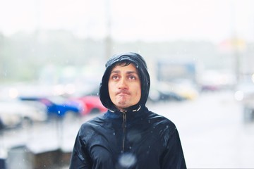Portrait of a young man in a jacket with a hood in the rain on blurred background city street,...