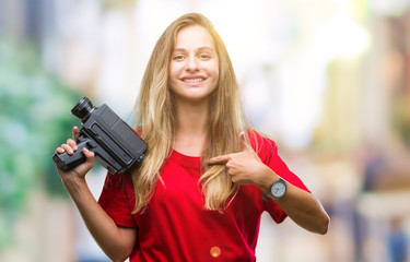 Young beautiful blonde woman filming using vintage camera over isolated background with surprise face pointing finger to himself