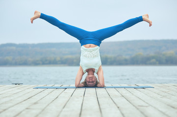 Beautiful young woman practices yoga asana Shirshasana - Headstand pose on the wooden deck near the...