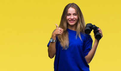 Young beautiful blonde woman looking through binoculars over isolated background happy with big smile doing ok sign, thumb up with fingers, excellent sign