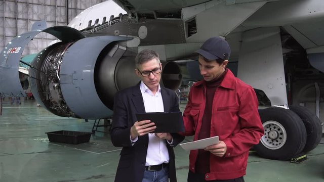 In a Hangar Aircraft Maintenance Engineer Shows Technical Data on Tablet Computer to Airplane Technician, They Diagnose Jet Engine Through Open Hatch.. 4K