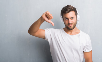 Handsome young man over grey grunge wall wearing headphones listening to music with angry face, negative sign showing dislike with thumbs down, rejection concept