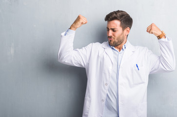 Handsome young professional man over grey grunge wall wearing white coat showing arms muscles smiling proud. Fitness concept.