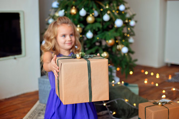 Obraz na płótnie Canvas Little beautiful girl with blond curly hair stretches a Christmas gift against the background of the Christmas tree. Christmas gift, New Year's gift Christmas concept.