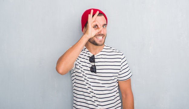 Handsome young hipster man over grey grunge wall wearing navy t-shirt and wool cap doing ok gesture with hand smiling, eye looking through fingers with happy face.