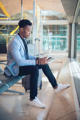 Young businessman seated in the airport checking a tablet and carrying a suitcase waiting for his flight