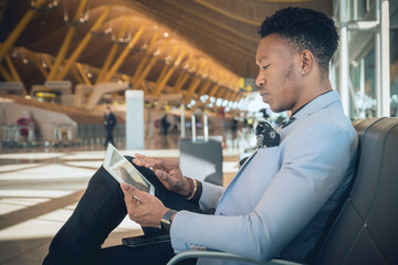 Young businessman seated in the airport checking a tablet and carrying a suitcase waiting for his flight