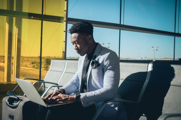 Young businessman is seated in the airport working with a laptop and carrying a suitcase waiting for his flight