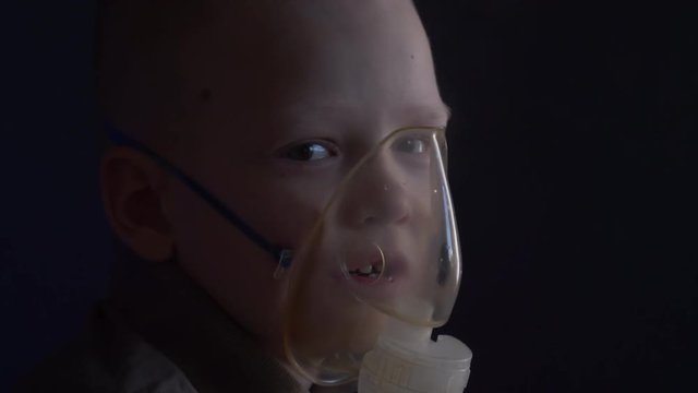 Unhappy boy with shaved head inhales vapors containing medicines for inhaler to stop cough. Child is looking information about his illness in smartphone.  Respiratory medicine. Bronchitis problem