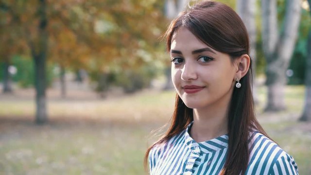 Three quarter front of romantic girl with shining eyes and long loose hair in striped dress standing with a happy and toothy smile in park in summer.