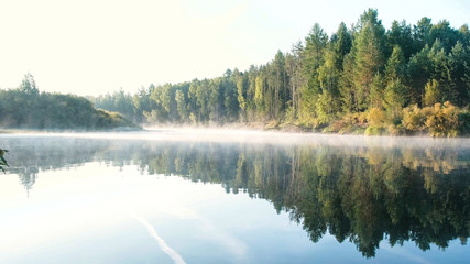 Fototapeta premium Mist over the river. Autumn forest is reflected in the water on the riverbank.