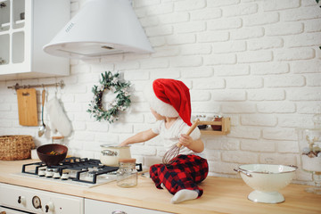 Cute little girl in santa hat, preparing cookies in the kitchen at home. Sits on the kitchen table and helps mom prepare a festive Christmas dinner