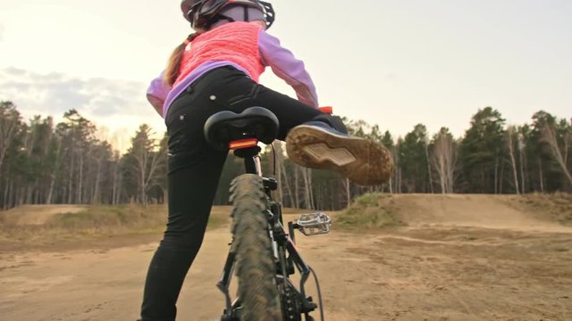 One caucasian children rides bike road track in dirt park. Girl riding black orange cycle in racetrack. Kid goes do bicycle sports. Biker motion ride with backpack and helmet. Mountain bike hardtail.