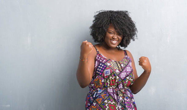 Young african american woman over grey grunge wall wearing colorful dress very happy and excited doing winner gesture with arms raised, smiling and screaming for success. Celebration concept.