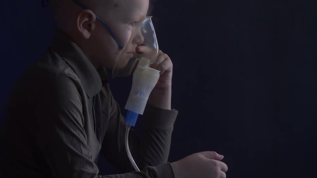 Unhappy boy with shaved head inhales vapors containing medicines for inhaler to stop cough. Child is looking information about his illness in smartphone.  Respiratory medicine. Bronchitis problem