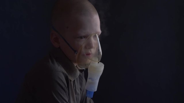 Unhappy boy with shaved head inhales vapors containing medicines for inhaler to stop cough. Child is looking information about his illness in smartphone.  Respiratory medicine. Bronchitis problem