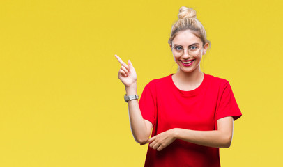 Young beautiful blonde woman wearing red t-shirt and glasses over isolated background with a big smile on face, pointing with hand and finger to the side looking at the camera.