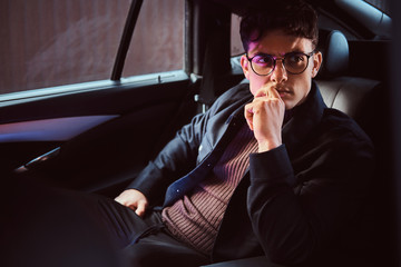 A pensive young man wearing glasses sitting in the back seat of the car.