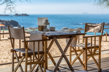 View of table and chairs in wood, with decorative elements and plate of reserve, beach club, sea as background