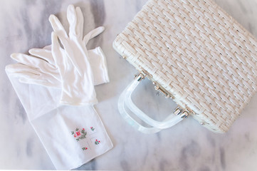 White Wicker Basket Purse with White Gloves and Handkerchief on a marble table