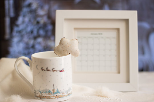 Close-up: mug and Christmas gingerbread on a white winter scarf. A blurred image of frame calendar is also on the table against background of a beautiful festive room with a decorated Christmas tree.