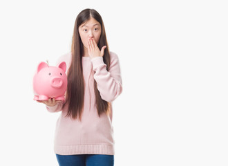 Young Chinese woman over isolated background holding piggy bank cover mouth with hand shocked with shame for mistake, expression of fear, scared in silence, secret concept