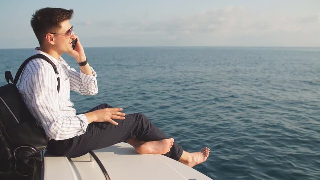 Space for text. Handsome young man with racksack in hands, dressed in office clothes relaxing and working on digital tablet on motorboat bow, over sea background