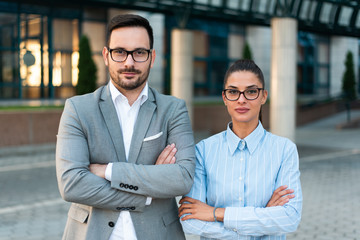 Young confident successful business people smiling, businessman and businesswoman standing together side by side as a team looking at the camera with crossed hands. Success concept.