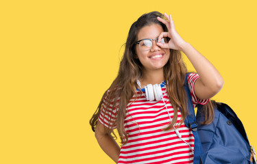 Young beautiful brunette student woman wearing headphones and backpack over isolated background doing ok gesture with hand smiling, eye looking through fingers with happy face.