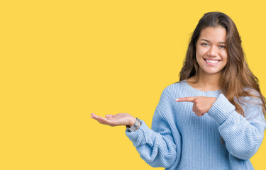 Young beautiful brunette woman wearing blue winter sweater over isolated background amazed and smiling to the camera while presenting with hand and pointing with finger.