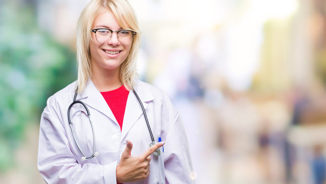 Young beautiful blonde doctor woman wearing medical uniform over isolated background cheerful with a smile of face pointing with hand and finger up to the side with happy and natural