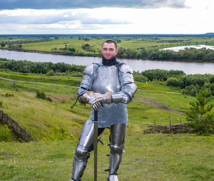 knight in armor stands against the background of native open spaces, forests and a river. Knightly armor and weapon. Semi - antique photo.