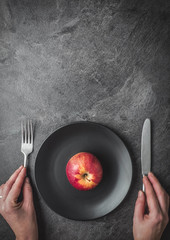 Red apple on black plate with knife and fork in female hands. Dark background. Overhead shot.