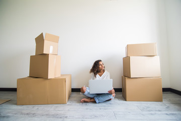 Attractive young latin american woman is moving, sitting among cardboard boxes, using a laptop and smiling