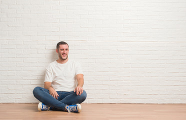 Young caucasian man sitting on the floor over white brick wall winking looking at the camera with...